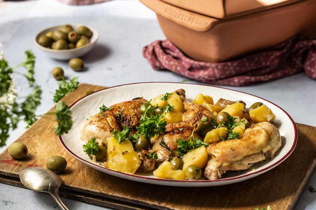 Teller mit gebratenem Hähnchen, Kartoffeln, Oliven und Petersilie auf Holzbrett. Schüssel mit Oliven im Hintergrund.