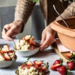 Person serviert Dessertschalen mit Streuseln und frischen Erdbeeren, dekoriert mit blühenden Zweigen auf einem Tisch.