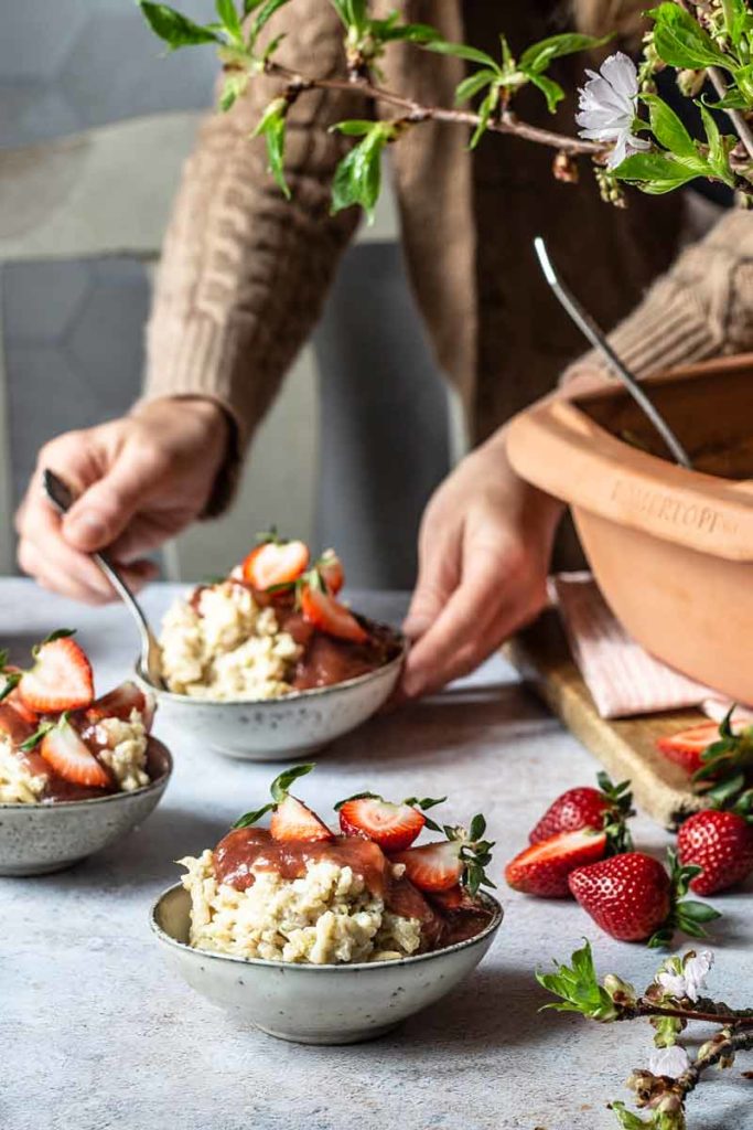Person serviert Dessertschalen mit Streuseln und frischen Erdbeeren, dekoriert mit blühenden Zweigen auf einem Tisch.