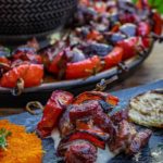 Gegrillter Fleischspieß auf Joghurtsoße mit Karottensalat und gegrilltem Gemüse, serviert auf einem dunklen Teller.