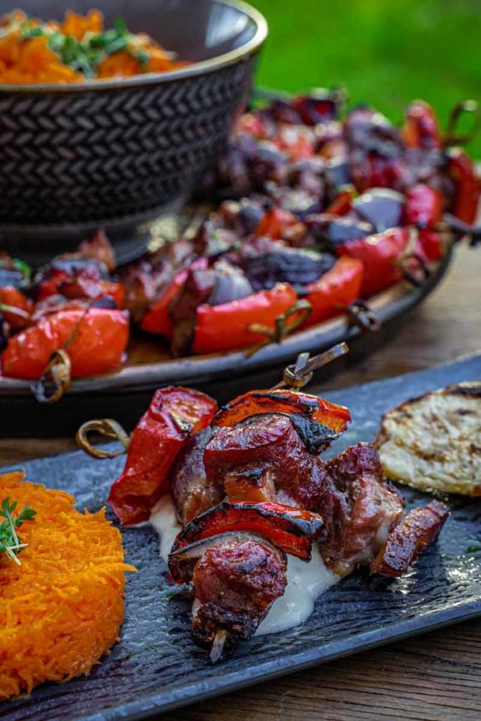 Gegrillter Fleischspieß auf Joghurtsoße mit Karottensalat und gegrilltem Gemüse, serviert auf einem dunklen Teller.