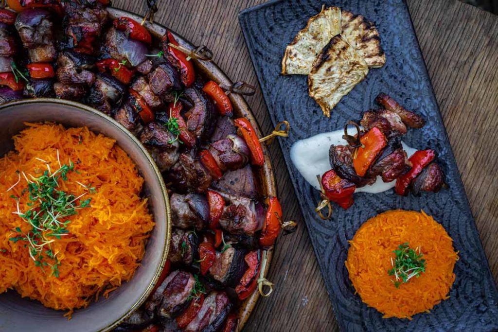 Bunte Grillspieße mit Paprika, dazu Karottensalat und gegrillter Sellerie auf Holzbrett serviert.