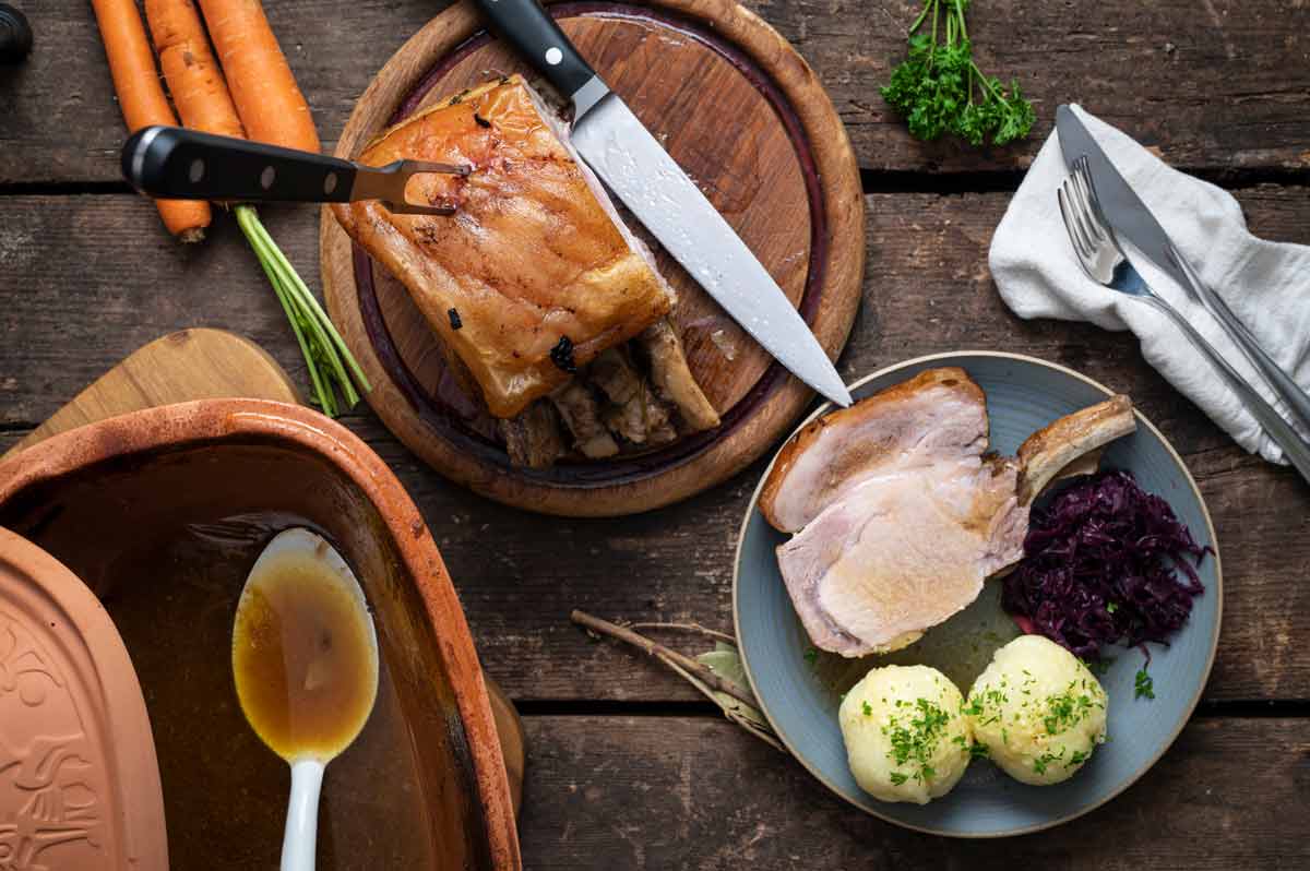 Braten auf Holzbrett und Teller mit Schweinebraten, Rotkohl und Klößen. Gemüse und Sauce im Hintergrund.
