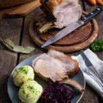 Teller mit Schweinebraten, Klößen und Rotkohl auf Holztisch. Im Hintergrund ein angeschnittenes Bratenstück mit Messer.