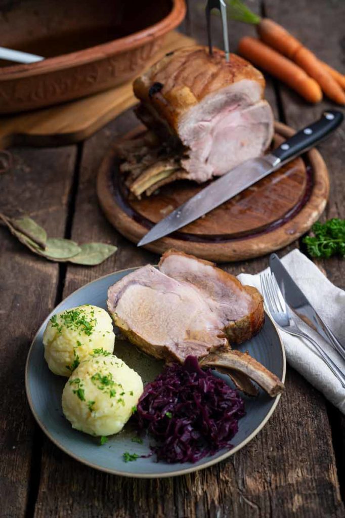 Teller mit Schweinebraten, Klößen und Rotkohl auf Holztisch. Im Hintergrund ein angeschnittenes Bratenstück mit Messer.