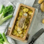 Gebackenes Fischfilet mit Kartoffeln und Fenchel in einer Auflaufform, daneben frischer Fenchel und Kartoffeln.
