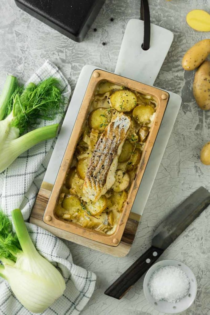 Gebackenes Fischfilet mit Kartoffeln und Fenchel in einer Auflaufform, daneben frischer Fenchel und Kartoffeln.