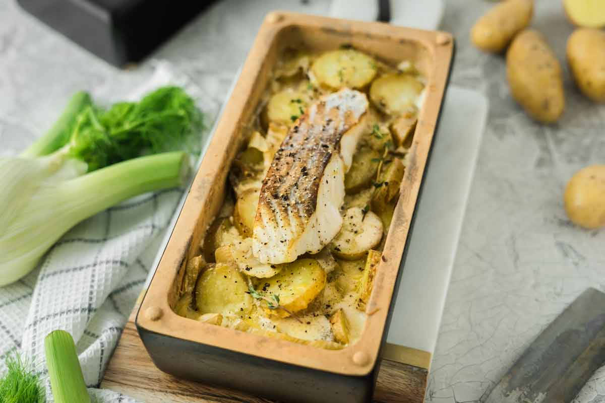 Gebratener Fisch auf Kartoffelscheiben in einer rechteckigen Tonform mit Kräutern, daneben rohe Fenchelknollen.