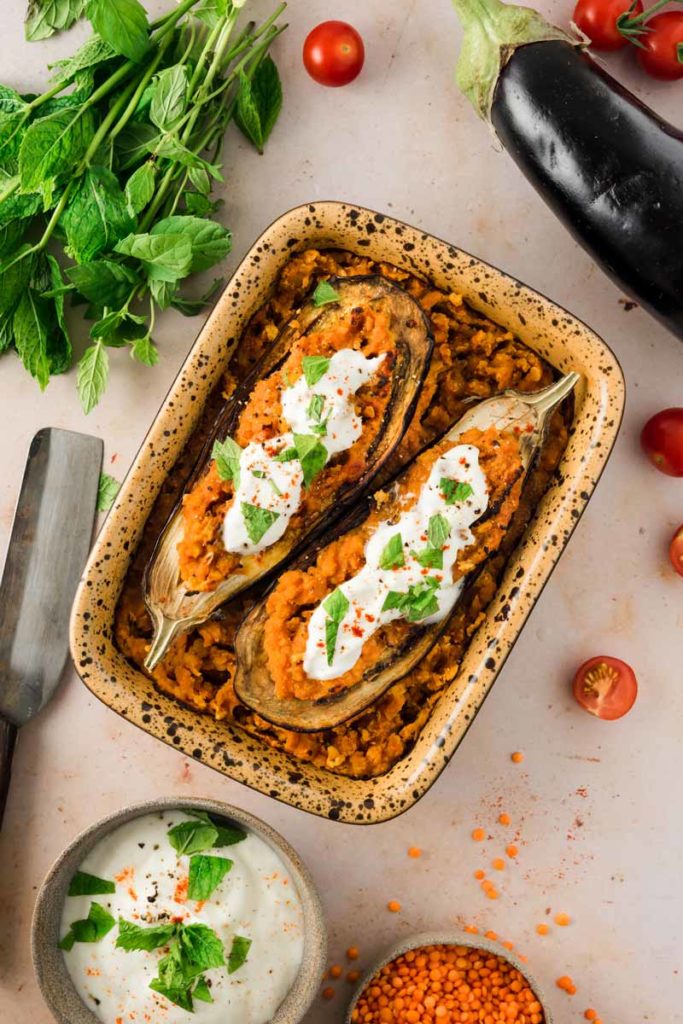 Gefüllte Auberginen mit roten Linsen, Joghurt und Kräutern in einer Auflaufform, dekoriert mit Tomaten und frischen Kräutern.