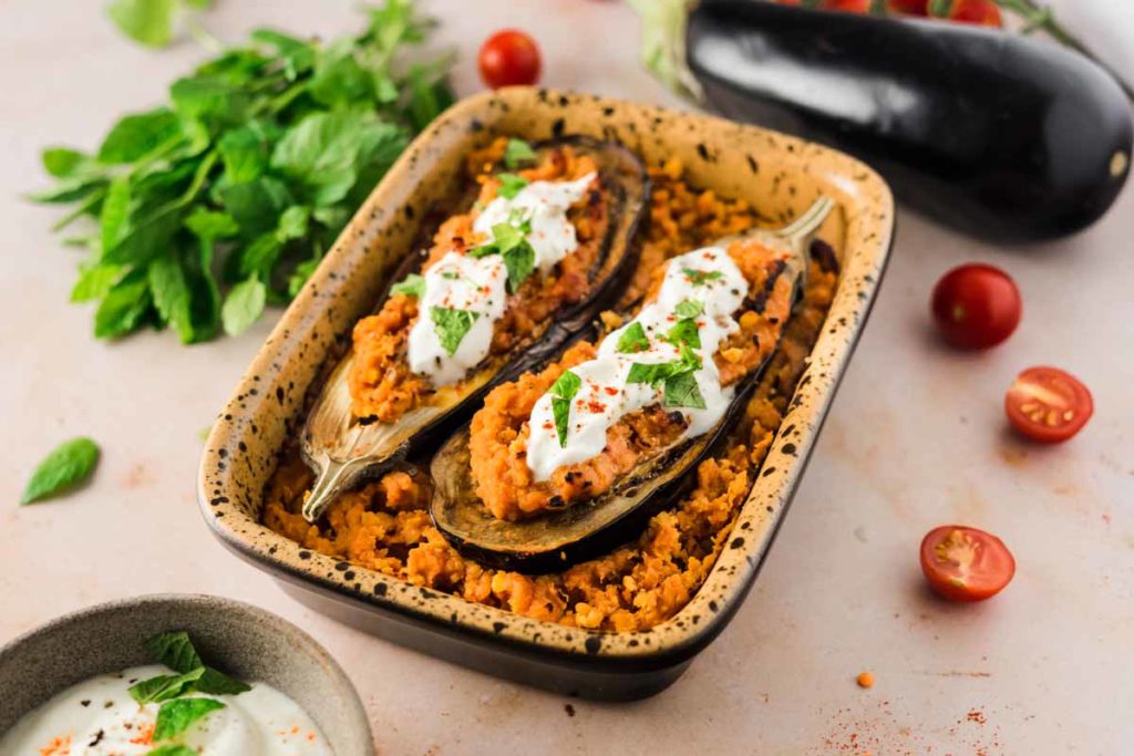 Gefüllte Auberginen mit Tomatensoße und Joghurt, garniert mit frischen Kräutern, in einer Keramikform, daneben Tomaten.