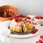 Gugelhupf mit Puderzuckerglasur und roten Johannisbeeren auf einem Teller, im Hintergrund eine Backform.