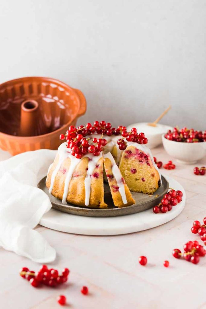 Gugelhupf mit Puderzuckerglasur und roten Johannisbeeren auf einem Teller, im Hintergrund eine Backform.