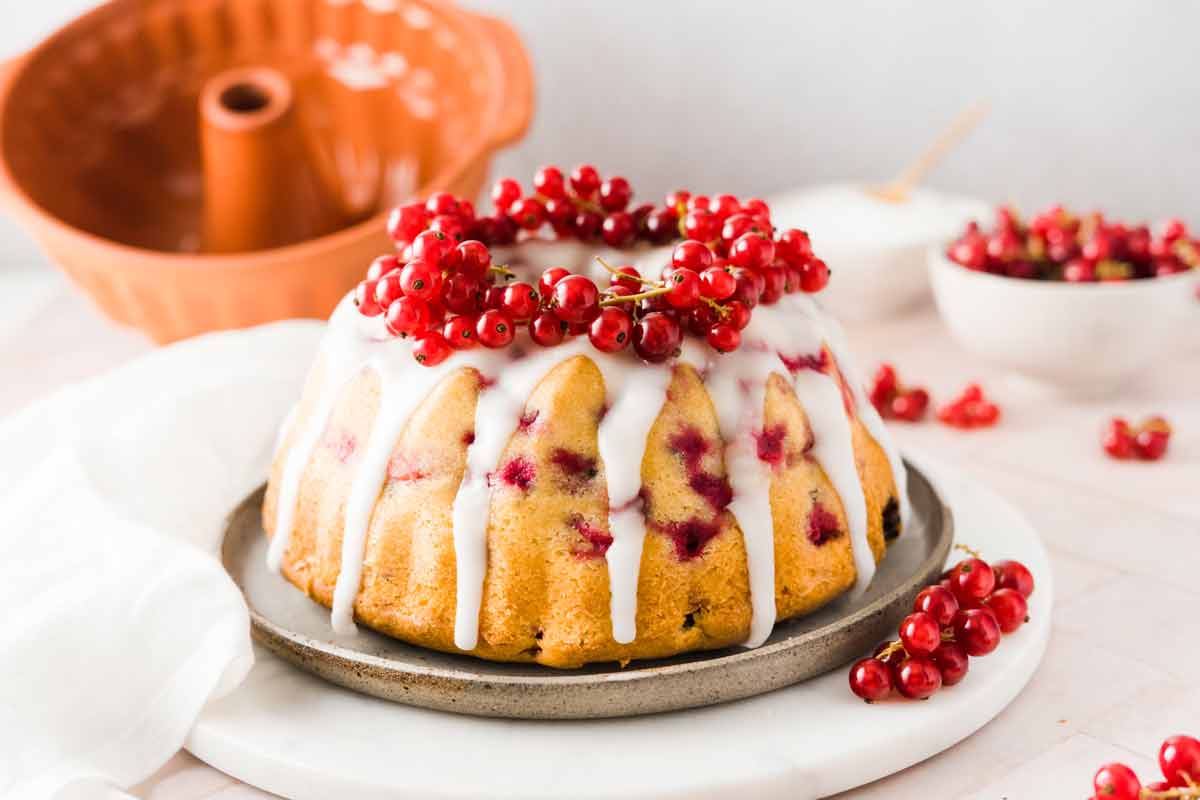 Gugelhupf mit Johannisbeerglasur, dekoriert mit roten Johannisbeeren auf Keramikteller. Hintergrund: Gugelhupfform, Johannisbeeren.