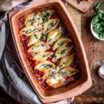 Gefüllte Muschelnudeln in einer Auflaufform auf Tomatensoße, garniert mit Käse und Basilikum. Hände halten die Form.