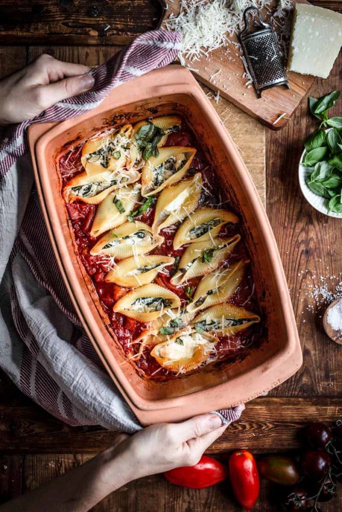Gefüllte Muschelnudeln in einer Auflaufform auf Tomatensoße, garniert mit Käse und Basilikum. Hände halten die Form.