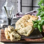 Frisch gebackenes Brot auf Gitterrost mit Kräutern, Salz und Besteck im Hintergrund, neben einem Basilikum.
