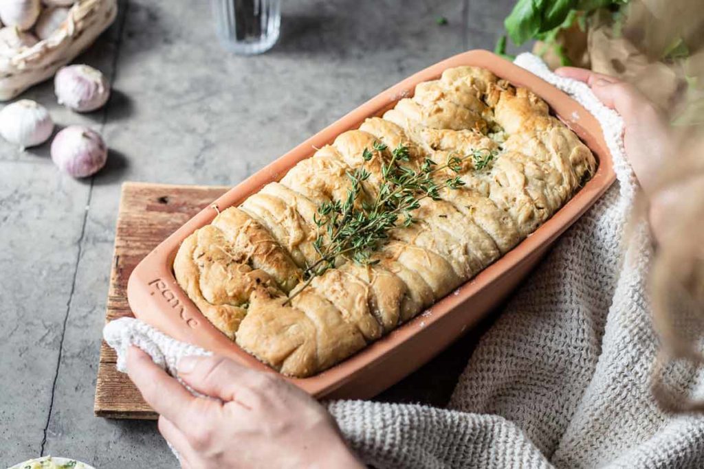 Frisch gebackenes Brot in einer Keramikform, dekoriert mit Thymianzweigen, wird auf einem Tisch serviert.