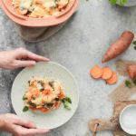 Hände servieren ein Stück Süßkartoffelauflauf auf einem Teller; daneben frische Kräuter und geschnittene Süßkartoffeln.