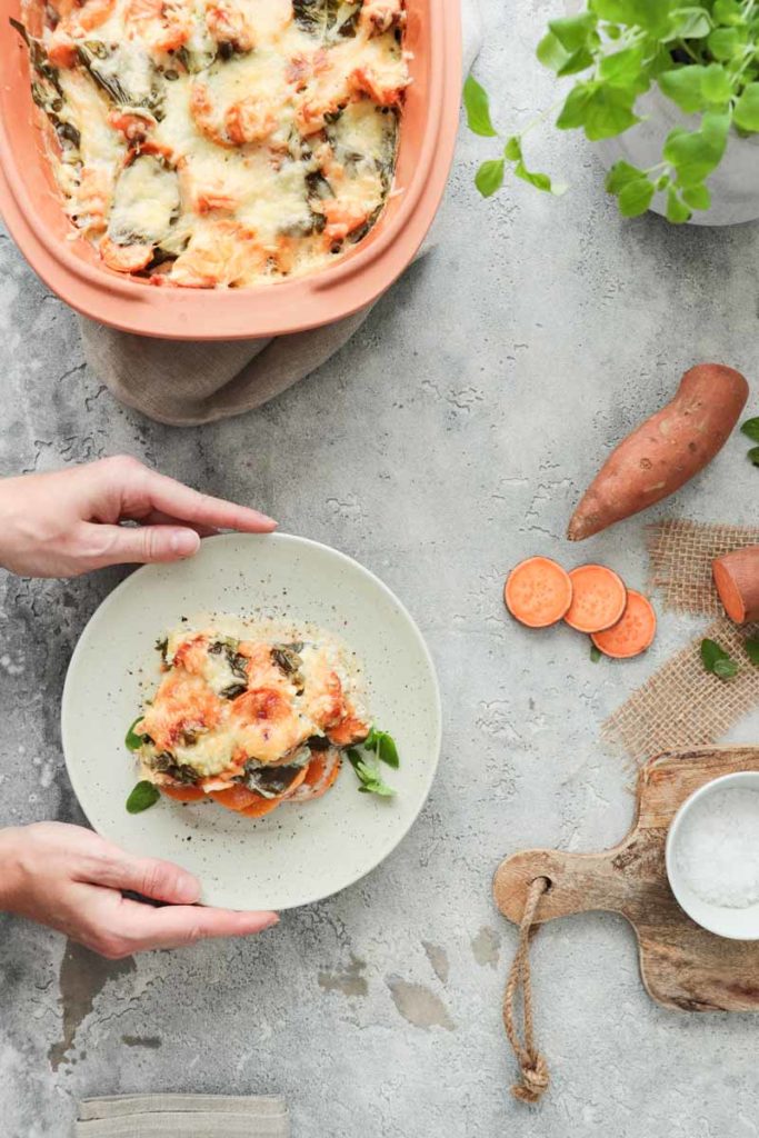 Hände servieren ein Stück Süßkartoffelauflauf auf einem Teller; daneben frische Kräuter und geschnittene Süßkartoffeln.