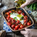 Hand gießt Öl über ofengebackene Tomaten und Feta in einer Backform, daneben Basilikum und Brot.