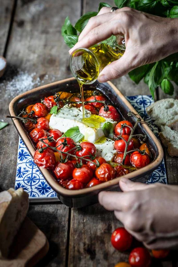 Hand gießt Öl über ofengebackene Tomaten und Feta in einer Backform, daneben Basilikum und Brot.