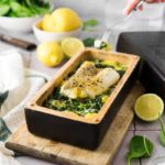 Gebackener Fisch auf Spinat in einer Kastenform, umgeben von Zitronen und einer Hand, die eine Gabel hält.