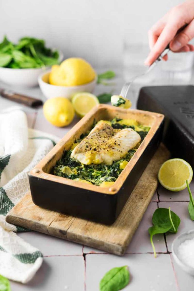 Gebackener Fisch auf Spinat in einer Kastenform, umgeben von Zitronen und einer Hand, die eine Gabel hält.