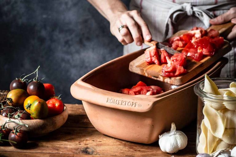Person schneidet Tomaten auf ein Holzbrett in einen Römertopf, neben einer Schüssel Tomaten, Knoblauch und Pasta.