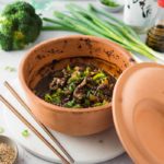 Ton-Gefäß mit Rindfleisch und Brokkoli, daneben asiatische Gewürze, Lauchzwiebeln und Stäbchen auf einem Marmorbrett.