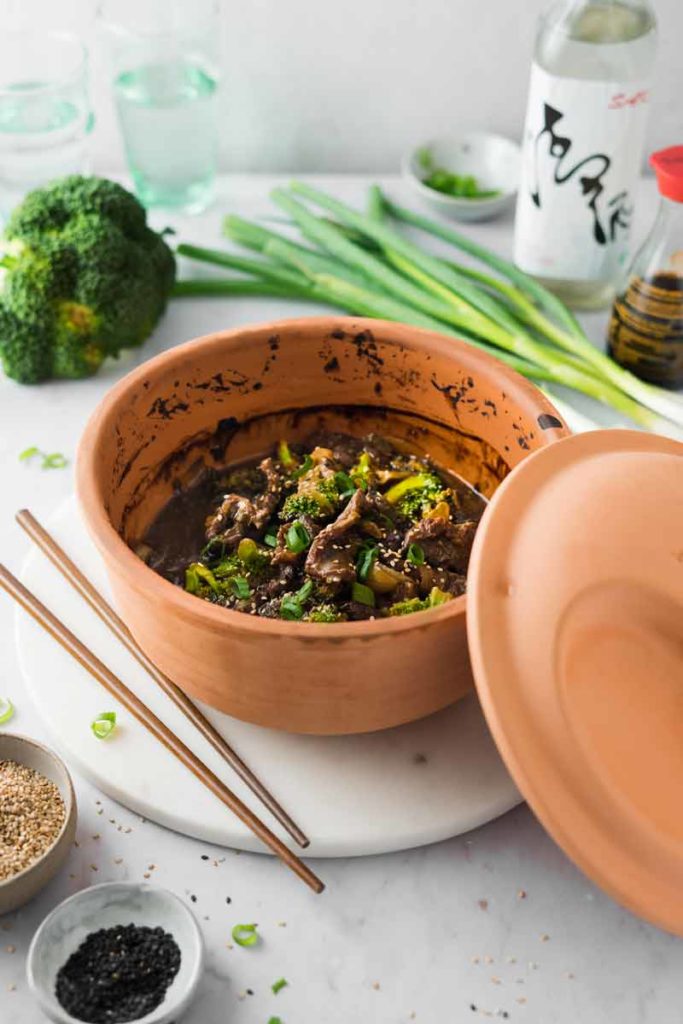 Ton-Gefäß mit Rindfleisch und Brokkoli, daneben asiatische Gewürze, Lauchzwiebeln und Stäbchen auf einem Marmorbrett.