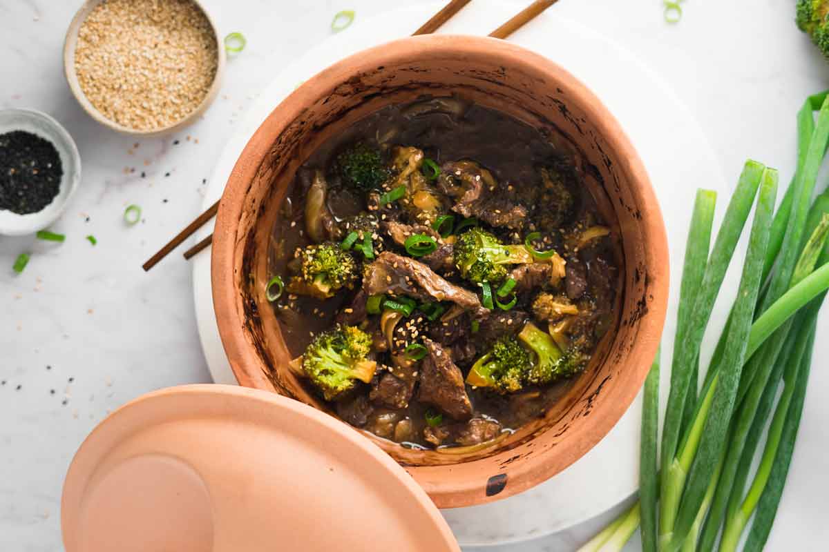 Eintopf mit Rindfleisch und Brokkoli in einem Tontopf. Daneben Frühlingszwiebeln und Schalen mit Sesam.