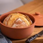 Frisch gebackenes Brot in einem runden, braunen Tontopf auf einem Holzbrett neben einem Brotmesser.