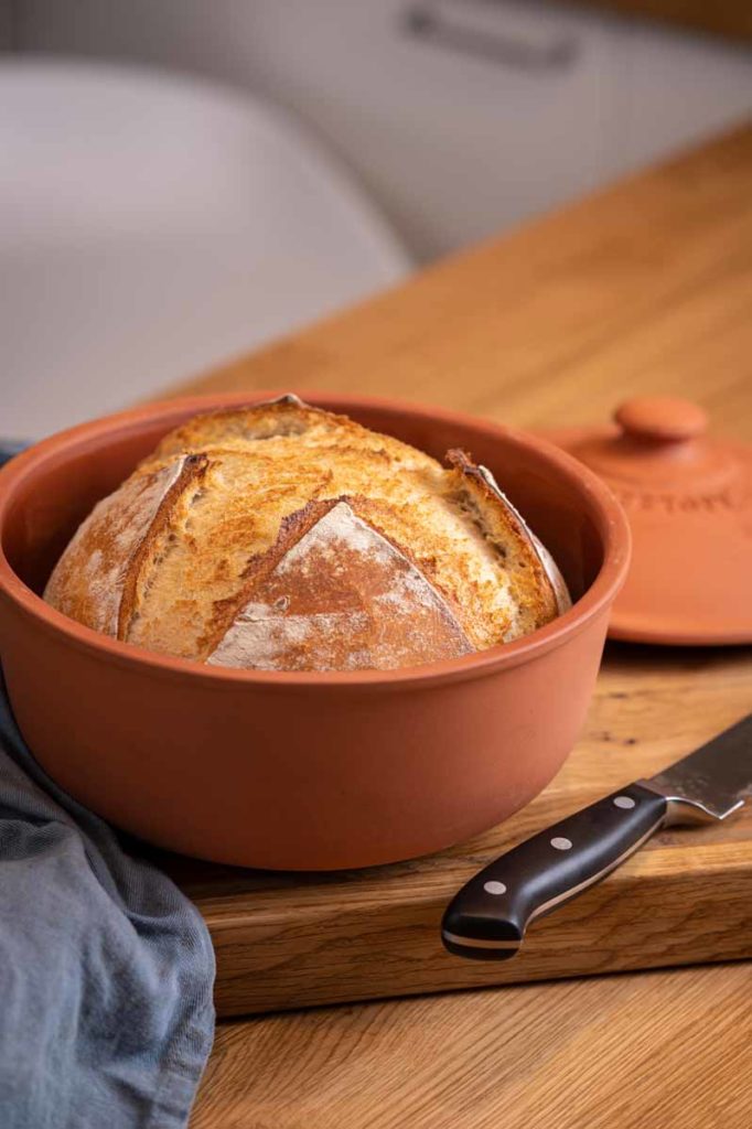 Frisch gebackenes Brot in einem runden, braunen Tontopf auf einem Holzbrett neben einem Brotmesser.