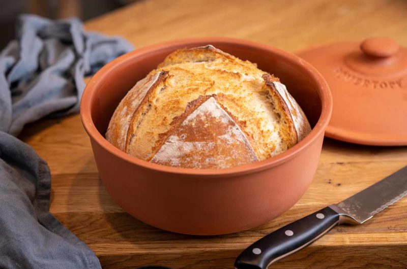 Rundes, goldbraunes Brot in einem Tontopf auf Holztisch, daneben ein Messer und ein blaues Tuch.