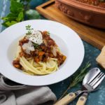 Teller mit Spaghetti, vegetarischer Bolognese, Ricotta und Kräutern, daneben Keramiktopf und Besteck auf blauem Tisch.