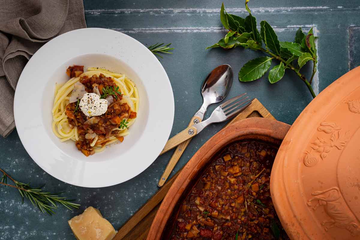 Spaghetti mit Gemüse-Ragout und Sahne in einer weißen Schale neben einem offenen Tontopf und Besteck.