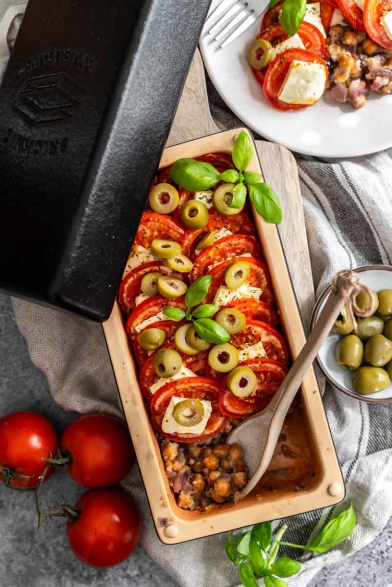 Ofenform mit Kichererbsen, Tomaten, Feta, Oliven und Basilikum. Daneben Tomaten und eine Schüssel mit Oliven.