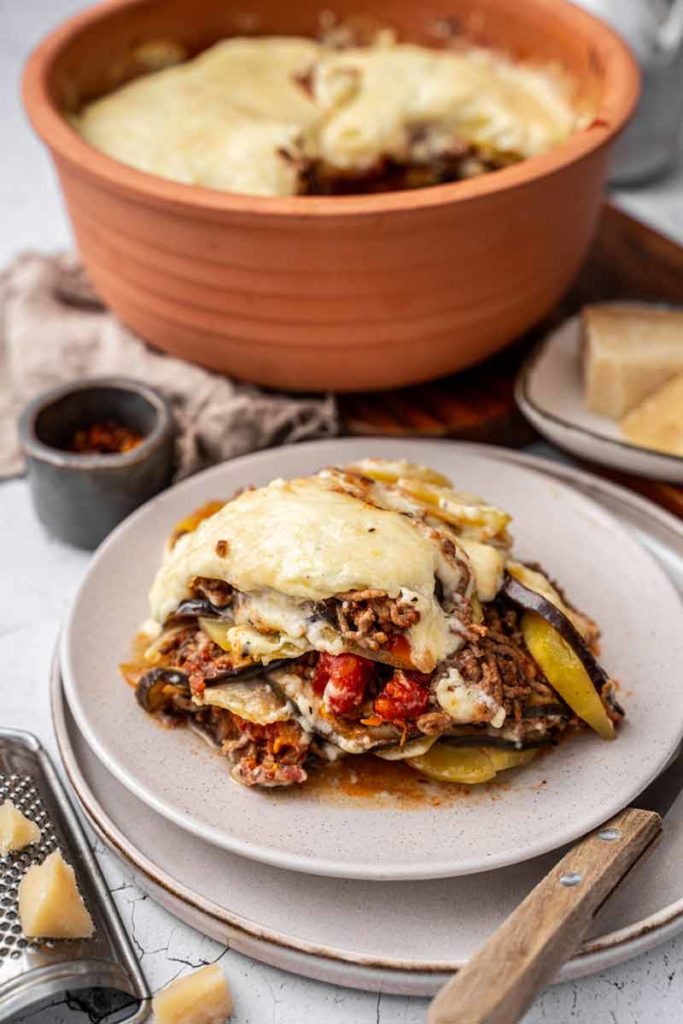 Servierraschen Moussaka mit Käse überbacken auf einem Teller vor einer Servierschale im Hintergrund.
