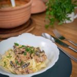 Weiße Schale mit Nudeln und Pilz-Sahne-Soße, garniert mit Petersilie. Im Hintergrund ein Topf und Kräuter.