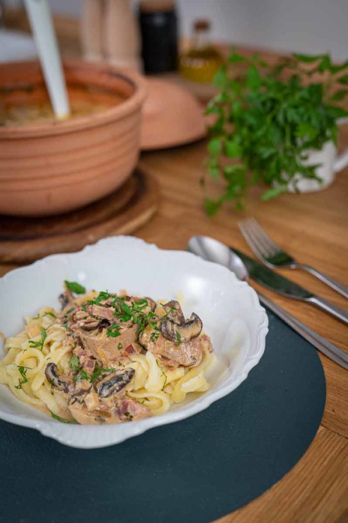 Weiße Schale mit Nudeln und Pilz-Sahne-Soße, garniert mit Petersilie. Im Hintergrund ein Topf und Kräuter.
