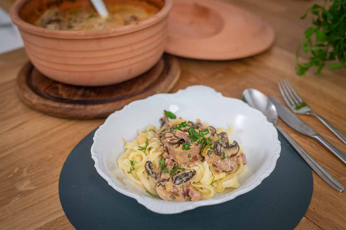 Weiße Schale mit Nudeln, Pilz-Sahnesauce, garniert mit Petersilie; daneben ein Terrakotta-Topf auf dem Tisch.