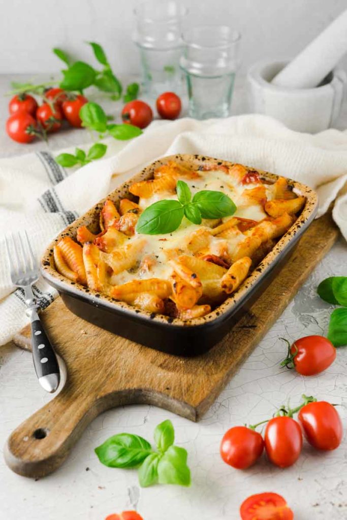 Gebackene Nudeln in Auflaufform mit geschmolzenem Käse und Basilikum, umgeben von frischen Tomaten und Basilikumblättern.