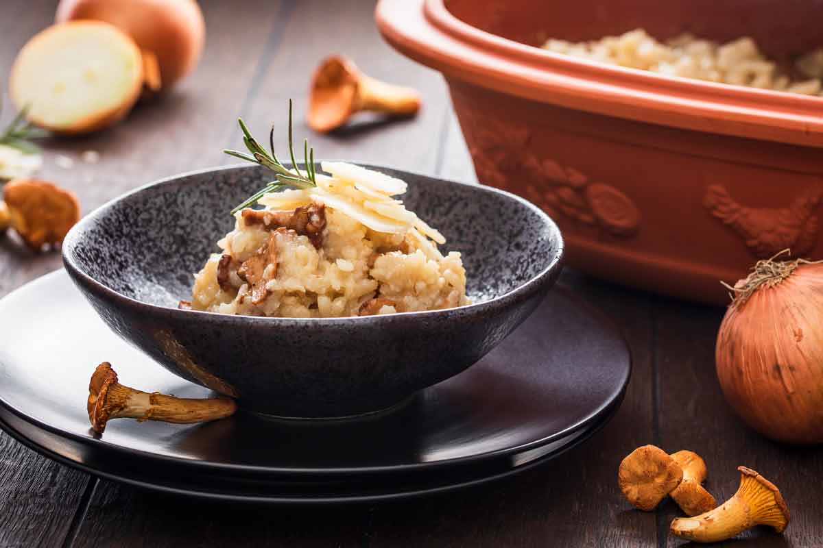 Schwarze Schale mit Pilzrisotto, garniert mit Rosmarin und Parmesan. Daneben Chanterellen und Zwiebel auf Holztisch.