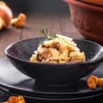 Risotto mit Pfifferlingen und Parmesanspänen, garniert mit einem Rosmarinzweig, serviert in einer schwarzen Schale.