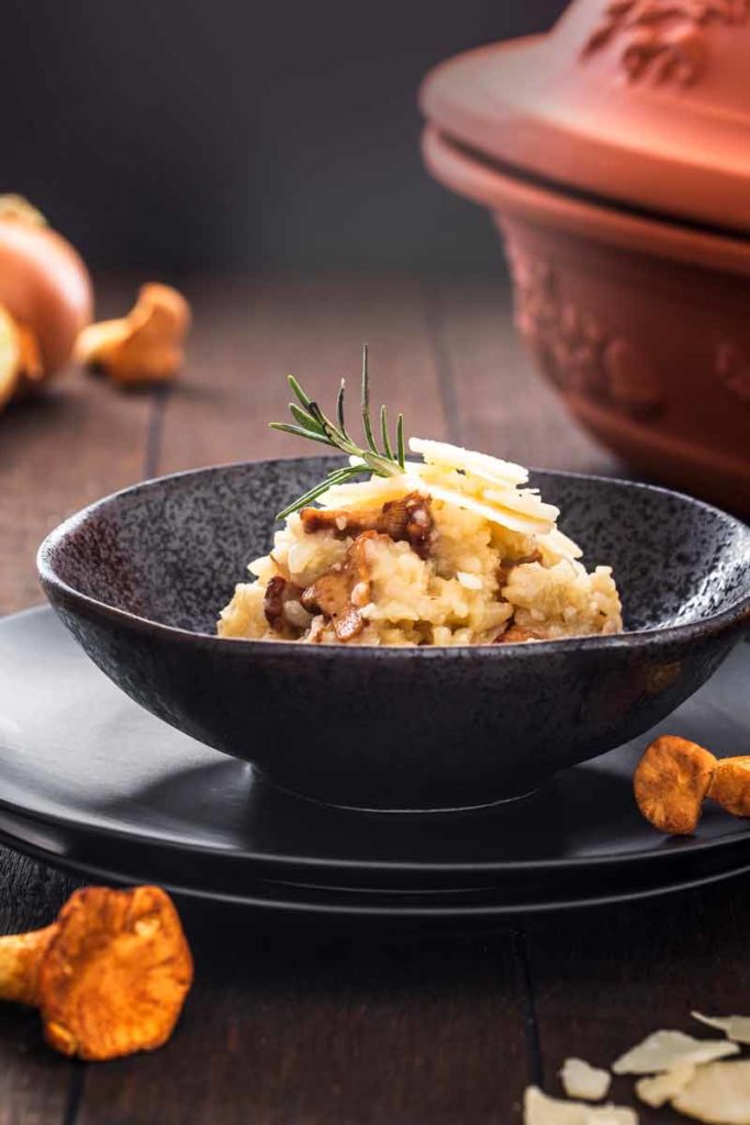Risotto mit Pfifferlingen und Parmesanspänen, garniert mit einem Rosmarinzweig, serviert in einer schwarzen Schale.