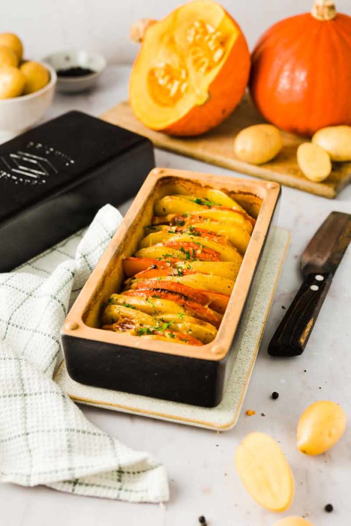 Herbstliches Gratin mit Kürbis und Kartoffeln in einer Auflaufform, umgeben von Kürbishälften und Kartoffeln.