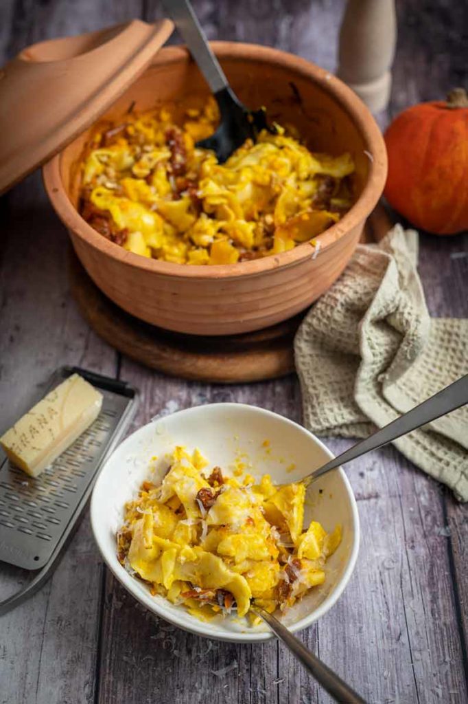 Ton-Auflaufform mit Kürbisnudeln, Schüssel mit Portion, Käsereibe, Parmesan, graues Tuch auf Holztisch.