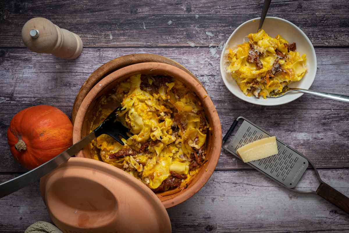 Ein Ton-Topf mit Kürbisnudeln, daneben ein Teller, eine Käsereibe und ein Kürbis auf Holztisch.