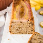 Bananenbrot auf einem Schneidebrett, dekoriert mit einer halbierten Banane, im Hintergrund liegen ganze Bananen.
