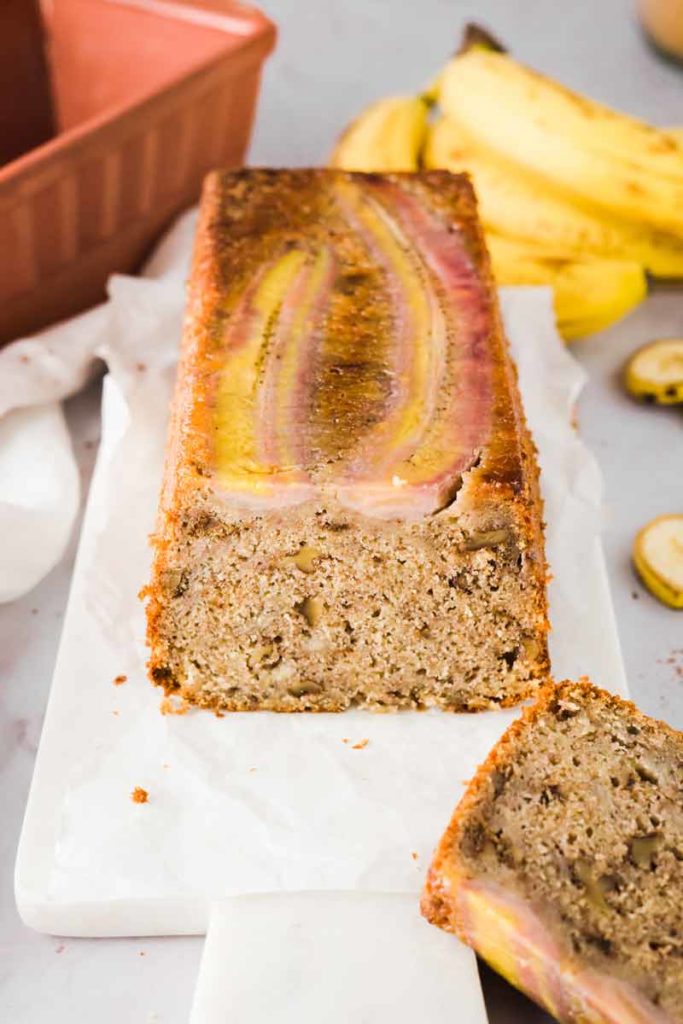 Bananenbrot auf einem Schneidebrett, dekoriert mit einer halbierten Banane, im Hintergrund liegen ganze Bananen.